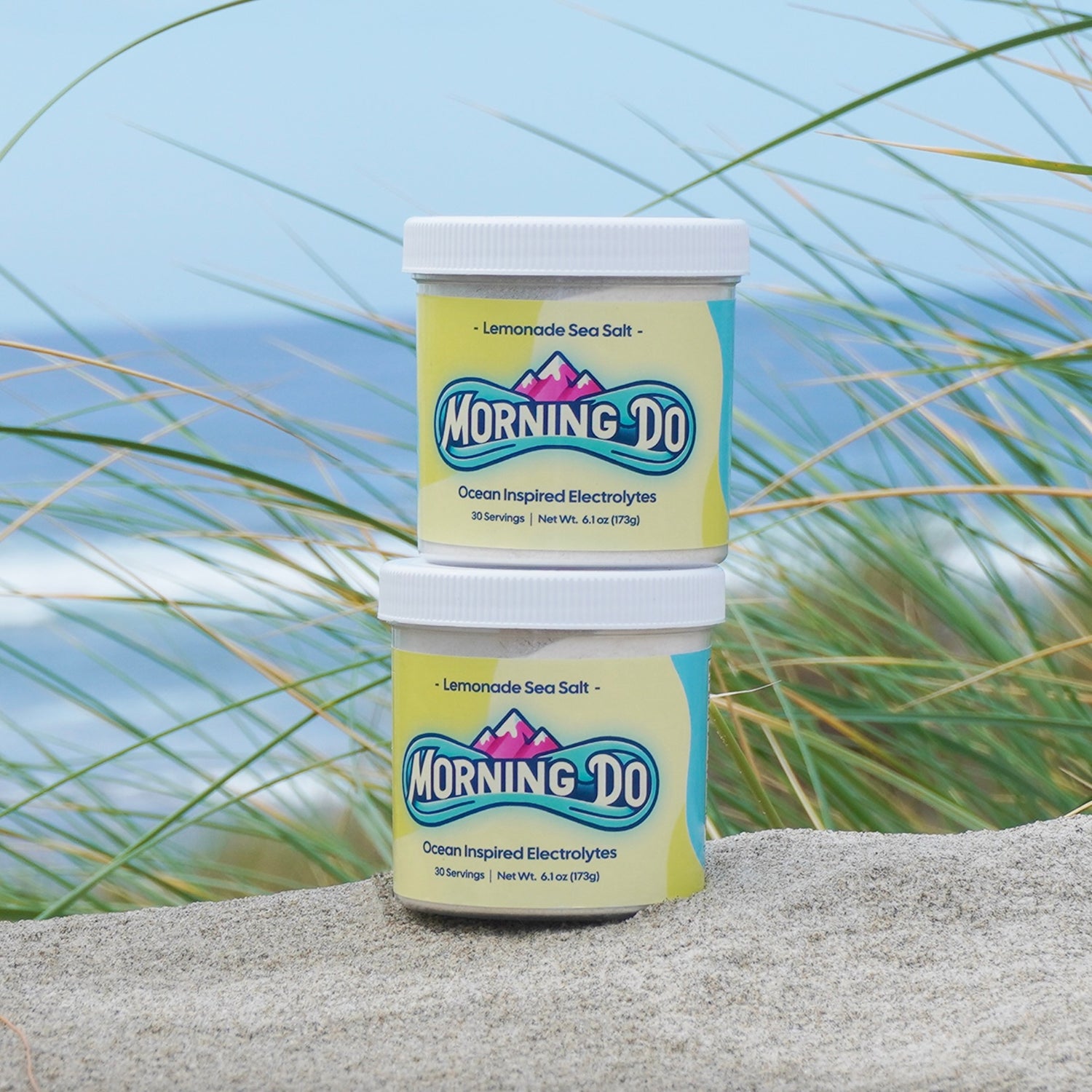 Morning Do Hydration Electrolyte Powder in Lemonade Sea Salt flavor displayed on a sandy beach with ocean and grass in the background. Featuring 30 servings of ocean-inspired electrolytes, this hydration drink supports replenishment and is perfect for maintaining hydration.